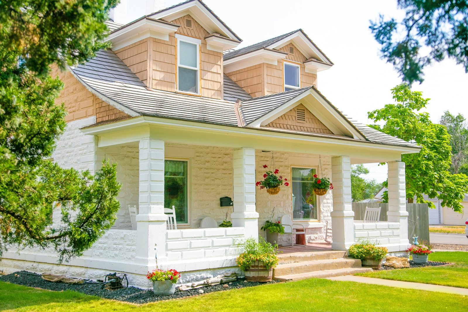 A charming two-story suburban house with light-colored stone and beige siding, featuring a welcoming front porch with white columns, hanging flower baskets, and potted plants. The well-maintained lawn and lush green trees frame the sunny, inviting exterior on a bright day.
